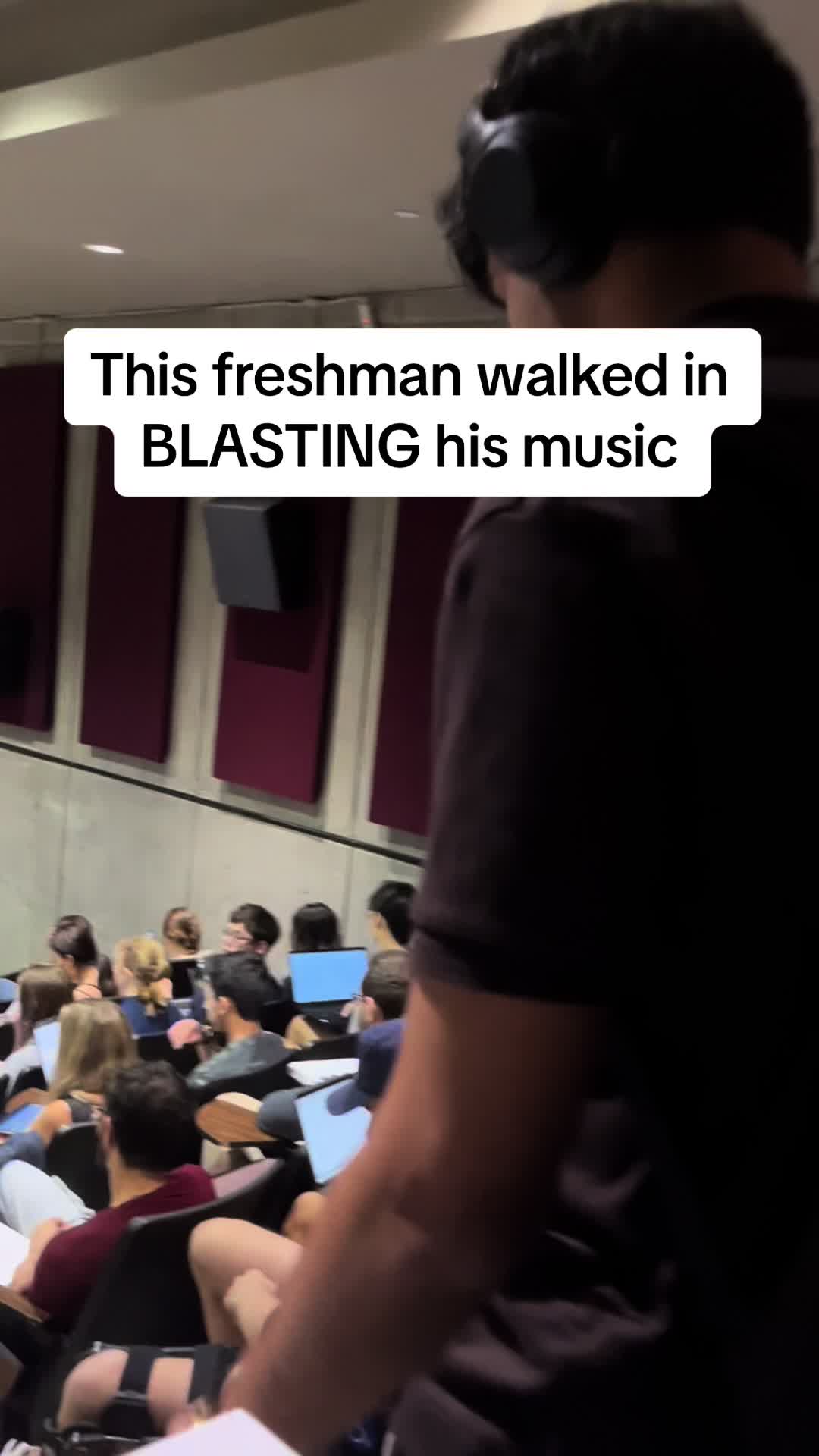 An image with a slightly obscured view of a college lecture hall. In the foreground, a person wearing headphones is partially visible from the back. The background shows rows of seated students with laptops in a classroom setting. Text in a white banner overlaying the image states: 'This freshman walked in BLASTING his music.' The lighting appears to be overhead classroom lights.
