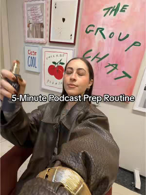 A woman is seated and spraying a bottle in front of her face. She is wearing a brown leather jacket and a gold belt. The background features framed artwork and text saying "The Group Chat". The image has a warm, inviting feel.
