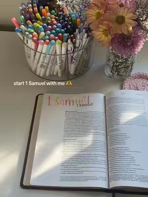 An open bible on a table, with the text '1 Samuel' highlighted in the image. Beside it, a jar of multiple colored markers and a vase of flowers.