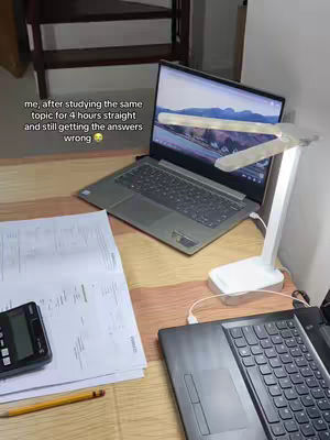 A top-down shot of a desk with a laptop, papers, and a calculator, lit by a desk lamp. The laptop screen displays a landscape scene. A pencil rests on the papers. Additional laptop and other study items are visible.