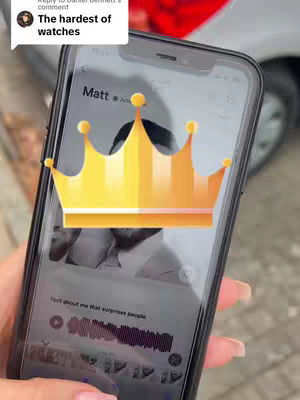 A hand holding a smartphone displaying an app screen. A golden crown graphic floats over the screen. In the top left is a profile picture and the text of a comment, while the background shows the exterior of a vehicle and road. Details are hard to discern due to blur.