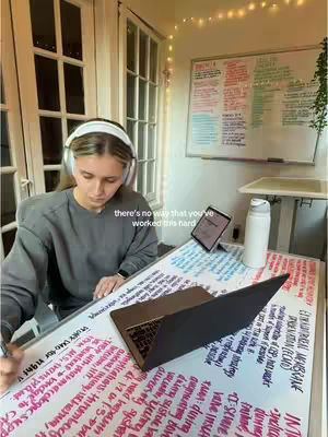 A young woman with blonde hair sits at a desk, studying. She wears headphones and is surrounded by study materials, including a laptop, tablet, and handwritten notes. The background features a whiteboard and a window, suggesting a home study environment.