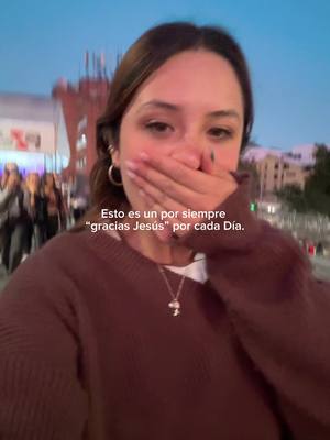 A young woman with brown hair is shown in a selfie, covering her mouth with her hand, with a city backdrop visible. The background is blurred lights and buildings, suggesting an outdoor evening setting. Text overlays the image, indicating thanks to Jesus for each day.