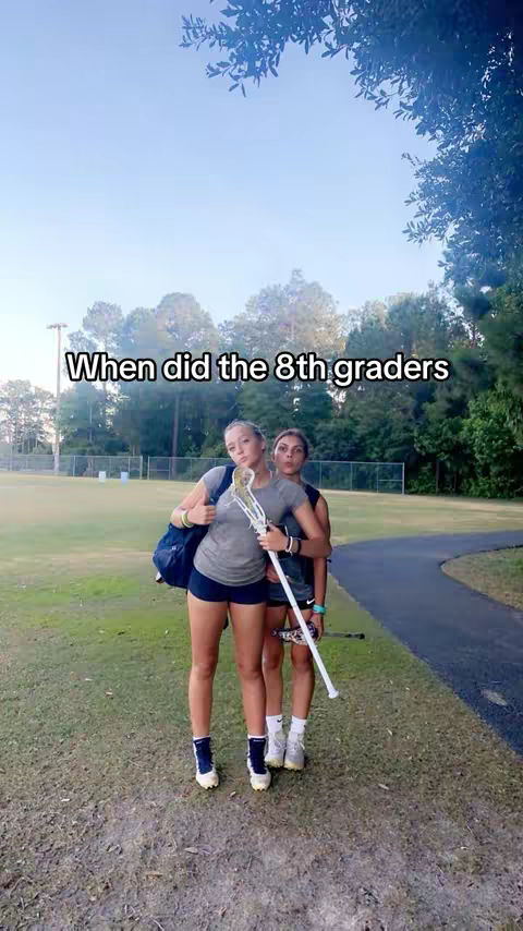 Two young women stand on a grassy field, likely a sports field, with one arm around the other. The woman on the left carries a lacrosse stick and a bag over her shoulder. They are standing in front of a field with trees in the background, with a hook text overlayed on the image.