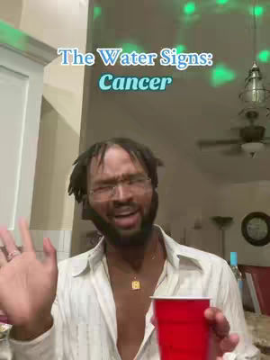 A man with glasses and a beard is holding a red cup while gesturing with his left hand. Text at the top says, 'The Water Signs: Cancer'. The background is a room with lights.