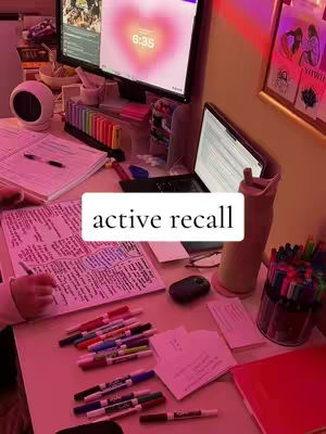 A well-lit desk setup with study materials, including notebooks, pens, and electronic devices. A text overlay indicates 'active recall.' The color scheme is warm, with pink and yellow hues.