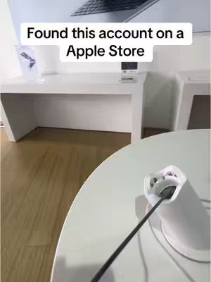 The image shows a modern, bright, minimalist interior of an Apple Store. A white table with a circular top is in the foreground, with a phone charging port and cable. In the background, there is a white desk, display shelves, and a monitor. The scene is brightly lit, with a slightly tilted perspective.