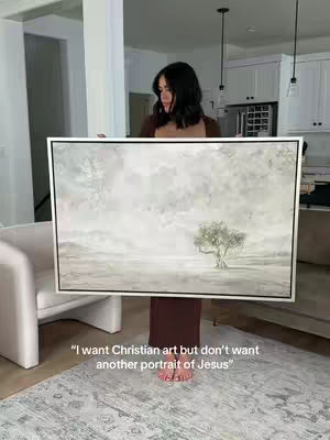 A woman is holding up a large framed canvas painting in a well-lit interior space. The painting has a muted, neutral color palette dominated by whites and grays and features an abstract landscape with possibly an olive tree. The woman is wearing a dark dress and standing on a rug. The background reveals a clean interior with white walls and cabinetry and some minimalist furniture.