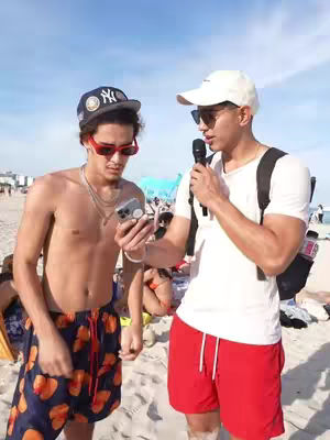 Two young men on a sunny beach. One is shirtless, wearing patterned swim trunks, sunglasses, and a baseball cap. The other wears a white t-shirt, red shorts, and a white baseball cap. They are standing close together on the sand talking, with a bright sky and beachgoers in the background.