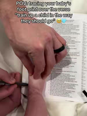 A person's hand is tracing a tiny baby foot on a white sheet with text on a paper. The image is shot from a first-person POV.