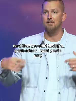 A medium shot features a man speaking in front of a soft purple and blue background, the man has short brown hair and is wearing a white undershirt and an open light denim collared shirt. Text overlayed on the image says 'next time you start having a panic attack I want you to pray'. He is gesturing with both hands, and there are microphones on the sides of his face showing he is giving a message.