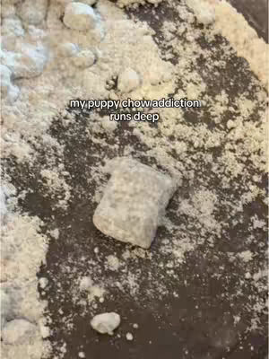 Close-up shot of what appears to be a bowl of 'puppy chow' snack mix. The composition is a bird's eye view, with the snack mix taking up the majority of the frame. The snack mix is composed of small, irregularly shaped, beige squares, likely cereal pieces, coated with a powdery, white substance, possibly powdered sugar or a similar flavoring. The pieces are scattered on a dark, irregular surface. Overlaid on the mix is the text "my puppy chow addiction runs deep."