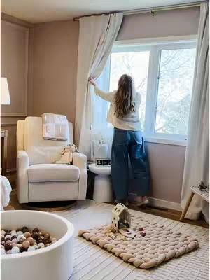 A woman stands near a window, pulling back a curtain to look out. She is dressed in a light-colored top and jeans. The room is decorated in a soft, neutral color palette, with cream-colored curtains, a beige rug, and a cozy armchair. In the foreground, there's a child's playroom with a ball pit and toy house, suggesting a nurturing domestic scene.