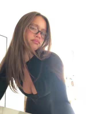 A woman with long brown hair and glasses looks down at the camera. She is wearing a black top and standing in front of a bright, white background.