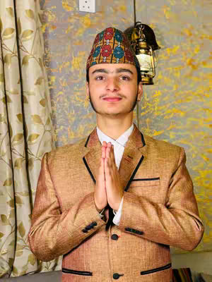 A young man wearing a traditional Nepali hat and a formal suit, with his hands pressed together in a prayer-like gesture. The background includes a patterned wall and a decorative lamp.