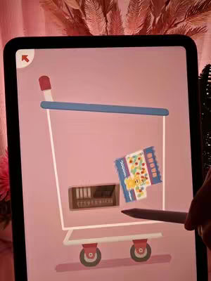 An iPad screen displays a cartoon shopping cart filled with items. The cart is white with red wheels and a blue handle. Inside, there's a box, and a packet of colorful items.