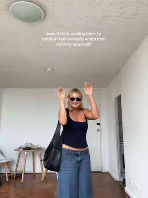 A woman with blonde hair and sunglasses is standing in what looks like an apartment with her arms raised in a greeting pose. She is wearing a black top and blue jeans. She has a black cross body bag slung over her shoulder. The background consists of white walls, a ceiling with a circular light, a small side table, and a doorway.