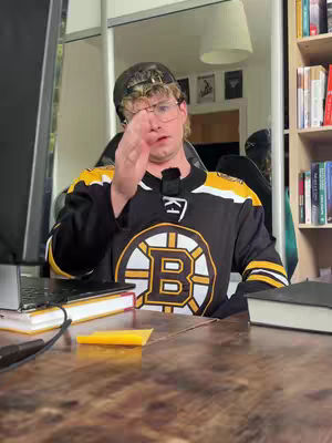 A man in a black and yellow hockey jersey gestures with his right hand towards the camera. He's wearing glasses and a black cap, with a small microphone clipped to his collar. A laptop and some books are visible on the wooden desk in front of him, and a shelf with more books can be seen in the background.