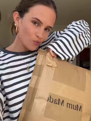 A young woman with light skin is angled toward the camera in a selfie. She has brown hair pulled back, gold hoop earrings, and a delicate gold necklace. She is wearing a black and white striped long-sleeved shirt and holding a large brown package up with both hands, which obscures the lower half of her body. Text that says "bsM mum" is visible on the package. The background is out of focus, with a neutral color palette.