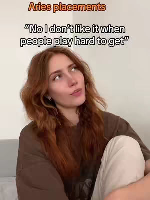 A woman with long, wavy red hair poses in a selfie-style shot, her upper body in view against a neutral background. She wears a brown shirt and appears to be seated, with her legs crossed and her expression open. Text overlays at the top of the image. The overall composition is well-lit, with a plain, uncluttered background that focuses attention on the woman and the text.