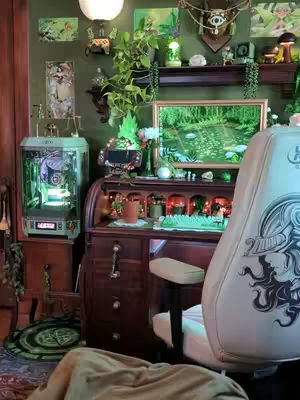 A cozy gaming setup featuring a wooden desk, a computer with a glowing case, and a comfortable chair. The room is decorated with plants, artwork, and warm lighting, creating a whimsical and inviting atmosphere.