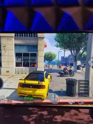 The image shows a screen capture of a video game featuring city scene with a yellow sports car parked on the side of a street with a building that has "Chappers" written on it. There is a person on a motorcycle and a trash can on the side of the road. The bottom of the screen shows the game's interface. The top has the image of the cover image being reflected on an object.