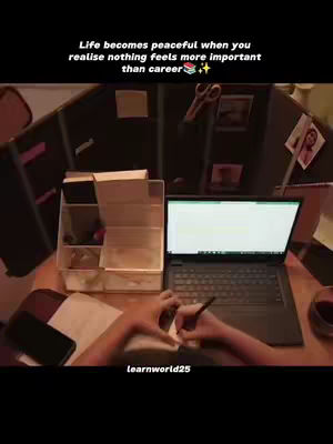 A desk with a laptop, a notebook, a smartphone, and a glass of liquid on it. A person is writing on the notebook. In the background, there are some shelves with stationary and a photo with female face.