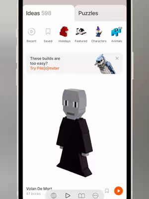 The cover image showcases an app interface on a smartphone, centered against a light grey background. At the bottom of the screen, a pixelated figure of Volan De Mort in a black robe stands out against a white background. Above him, various options are available, including 'Ideas', 'Puzzles', and tabs like 'Recent', 'Saved', 'Holidays', etc., with related icons. A pop-up notification reading, 'These builds are too easy? Try Pile[o]meter' is visible near the top. The layout is clean and minimalist, with most elements being simple, colored icons and text on a white backdrop.