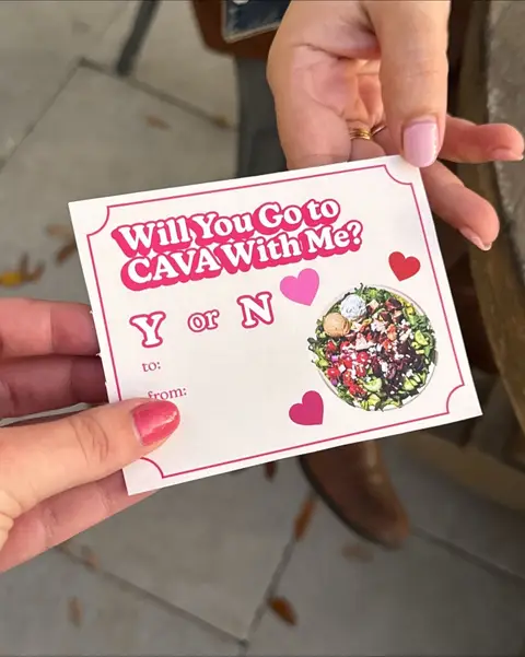 Two hands hold a white card with text and imagery related to CAVA restaurant. The card has a pink border and is being held against a blurred background. The card's text is in pink, with the words arranged to form a proposal.  A graphic of a salad bowl is shown on the card, along with several hearts.