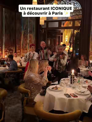 The image shows a restaurant interior with a festive atmosphere. The foreground features a table set for dining, complete with a white tablecloth, plates, glasses, and a lit candle. People are seated around the table, some looking toward a band playing music in the background. A group of musicians, including a violinist and an accordion player, are positioned in the center, adding to the celebratory mood. The decor includes ornate wall panels and chandeliers, with additional diners in the background.
