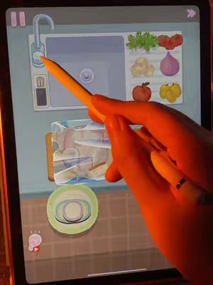 The image shows a person's hand holding an orange stylus, interacting with a tablet screen displaying a stylized cooking game interface. The screen shows a sink with vegetables and fruits, and a plate with food on it.