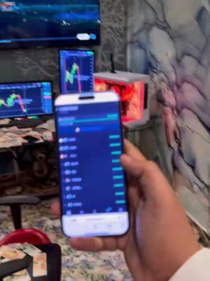A hand holding a smartphone displaying a trading app interface. Multiple computer screens with trading charts are visible in the background. The setting appears to be an office or a room dedicated to trading activities, with cables, a desk and money visible.