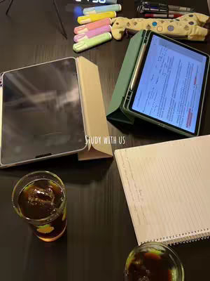 A top-down shot of a dark wooden surface with several items suggestive of a study session. Two tablets are present, one open with its screen visible, and the other closed. A notepad, several pens, and multiple colorful highlighters are laid out beside them. Additionally, there is a glass with a dark liquid and ice cubes, another glass visible at the bottom, and the top of an alarm clock are also in the frame.