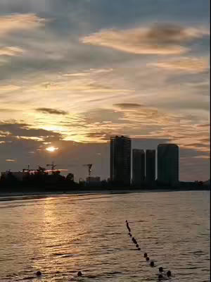 The image captures a serene sunrise over a body of water, likely a sea or large lake. Silhouetted against the bright orange and yellow hues of the rising sun are dark, tall buildings, perhaps a city skyline. The water reflects the light creating a shimmering effect. A line of buoys or other objects extends into the water adding some depth.