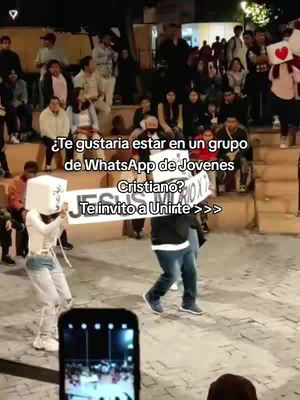 Crowd shot of a gathering of young people in an outdoor setting. In the foreground, two people hold a sign with text inviting viewers to join a WhatsApp group for Christian youth. The sign obscures the people's faces. Other people are visible in the background, some seated and others standing. The setting appears to be a public space, with stone steps and outdoor lighting. The scene is lit with natural light, and the overall composition suggests a community gathering.