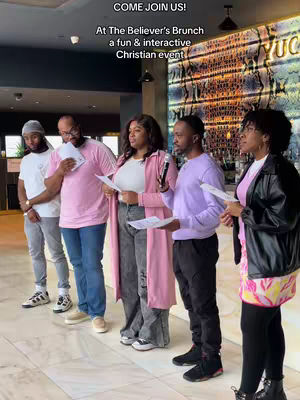 A group of diverse people stand in a well-lit space next to an illuminated wall display. They are dressed in a variety of colors, with a pink-themed outfit prominently displayed, holding papers and microphones, suggesting a presentation or event.