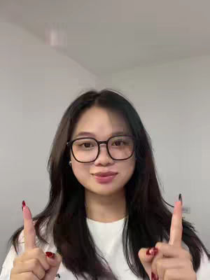 A woman with long dark hair and glasses is pointing upwards with both index fingers. She appears to be in a brightly lit room, with a plain background.