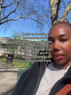 A selfie-style shot featuring a Black woman against a backdrop of a park with trees and a cloudy sky. On her face are two piercings on her nose and 1 on her lip. She's wearing a black hoodie, and earbuds. On the screen is a text overlay with an inspirational quote.