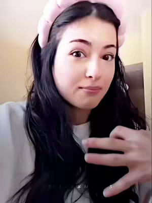 A selfie-style shot featuring a woman with dark hair pulled back into pigtails and a thick pink headband. She has a light complexion and is looking at the camera with a slight smile. Her right hand is up, fingers splayed, near her face. The background is softly lit with cream and tan walls.