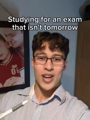 A young man with short, curly dark hair wearing glasses and braces, is centered in the frame, looking at the camera and speaking. He is wearing a light blue button-down shirt over a white undershirt and holding a pen. Behind him is a framed picture of other people, and a light blue wall on either side.