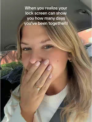 A selfie-style shot of a woman inside a car. She is looking towards the camera with a surprised or amused expression, her hand covering her mouth. The background is slightly blurred, suggesting movement or a shallow depth of field. The layout focuses on her face and upper body.