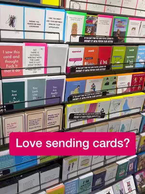 A retail display of various greeting cards with humorous and relatable messages. The foreground features a bright pink banner with a text overlay.