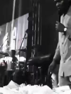 A black and white image shows a man in a light suit speaking into a microphone. He is standing on what appears to be a stage covered with scattered white objects, possibly paper. Behind him are musical instruments and other stage equipment against a dark background.