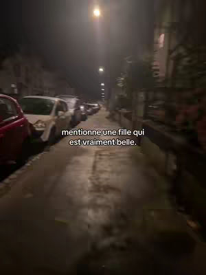 A low-angle shot taken at night, capturing a city street with parked cars on the right side and a wet, reflective pavement leading into the distance. Overhead, streetlights illuminate the scene, and text overlays are present.
