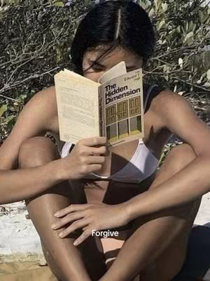 A woman sits cross-legged outdoors, partially submerged in a bright setting, while holding a hardcover book titled 'The Hidden Dimension' close to her face, with her eyes obscured. The background features lush green foliage.