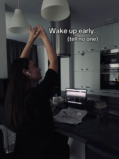 A young woman in a kitchen, illuminated by overhead lights. She's stretching with her arms raised, wearing earbuds, a laptop on the counter, and papers nearby, suggesting a study or work session. The scene is mostly dark, the lighting is soft and natural.