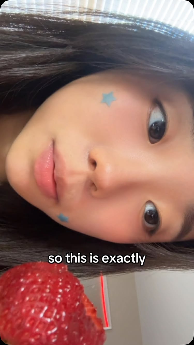 A selfie-style shot of a young person with dark hair, face angled slightly towards the camera. The person has light skin, with a pinkish tone to their cheeks, and full lips. There are two small, light blue star-shaped stickers applied on each side of the face, near the cheekbones. The person seems to be holding or near a cut strawberry in the bottom half of the shot. The hook, 'so this is exactly' is written in white letters on the image, near the face.