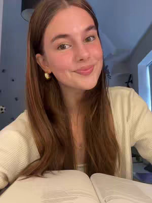 A young woman with long auburn hair and gold hoop earrings sits at a desk with a book open in front of her. She smiles gently, gazing directly at the camera. Her blouse is white, and the background is softly lit, suggesting a home study setting. A touch of blush highlights her cheeks, contributing to a warm overall composition.