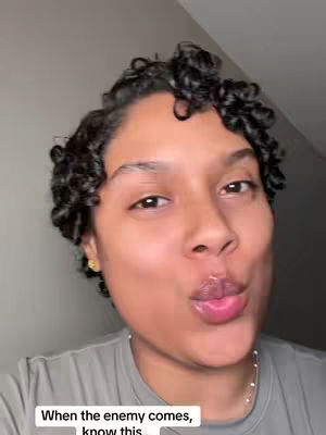 A close-up shot of a person with short, dark curly hair and fair skin, pursing their lips in a kissing expression. They are wearing a gray shirt and a delicate necklace, with a light gray backdrop and an overlay text at the bottom.
