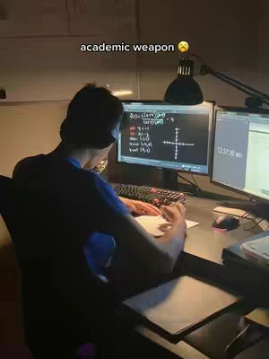 A person is seen from the back, sitting at a desk late at night. Two computer monitors are illuminating the workspace. The person is hunched over a sheet of paper with a pen in hand, taking notes or working on calculations. On the screen, equations and a graph are visible, suggesting that the topic is mathematics or calculus.